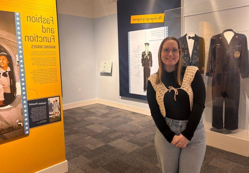 Juliette Good stands in front of glass cases holding uniforms and walls displaying information about the history of Navy uniforms.